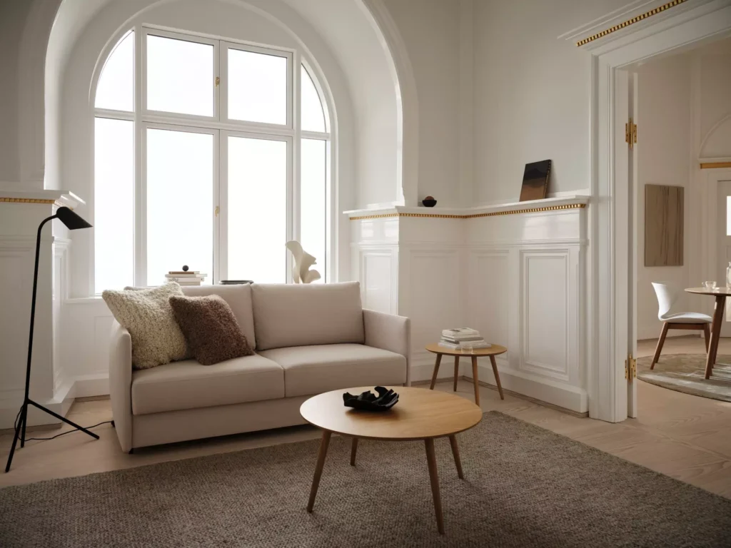 Jasna sofa skandynawska w eleganckim salonie z dużym oknem, drewnianym stolikiem i minimalistycznymi dodatkami