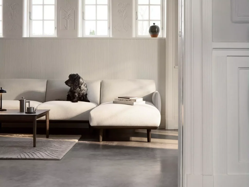 Sofa Aarhus BoConcept z szezlongiem, drewnianą podstawą i stolikiem kawowym w jasnym salonie inspirowanym skandynawskim designem.